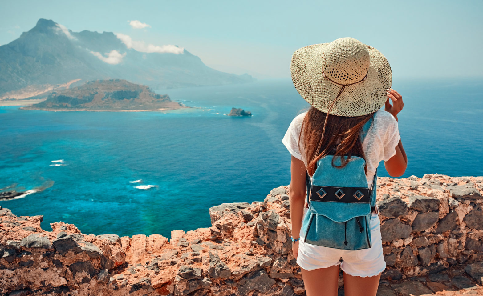 Une jeune femme en voyage portant un chapeau de paille et un sac à dos bleu turquoise contemple un paysage méditerranéen, avec vue sur la mer, les navires et les montagnes au loin